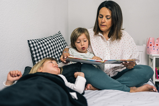 Mother sitting on the bed watching with her blonde son her favorite story while her other blonde daughter is lying on the bed watching them.