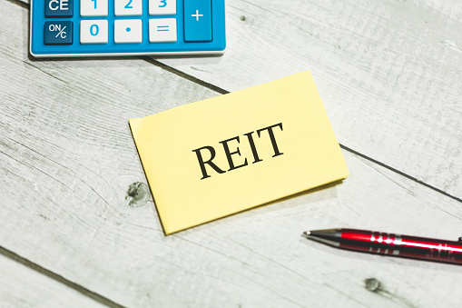 The acronym REIT for Real Estate Investment Trust written on a piece of paper lying on a wooden table. A calculator and a pen in the composition. REIT Concept.