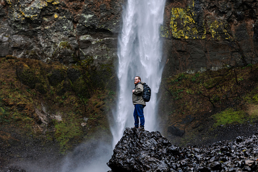 A caucasian male hiker in his 20s standing at the base of a large waterfall. Shot in the Columbia River Gorge in Oregon.