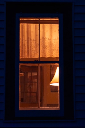 Softly lit, soft-focus window of a farmhouse, lit from within at night. Lampshade, thin curtains, warm amber glow. Closely framed. Painterly, vintage mood.
