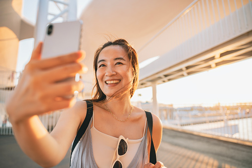 An Asian woman captures beautiful travel memories by taking selfies with her smartphone when she comes across stunning harbors and distant city architectures during her journey.