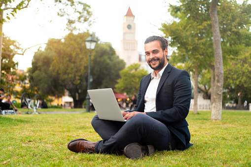 Handsome hispanic male businessman sitting lotus position in public park use laptop. Remote work concept. Stylish european business person in park summer day working on computer. Technology.