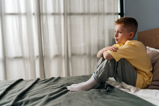 Side view of lonely sad little boy sitting on bed with hugging knee and sadness looking away, feeling pressure and problem. Thoughtful stressed child resting alone in bedroom with negative emotion.