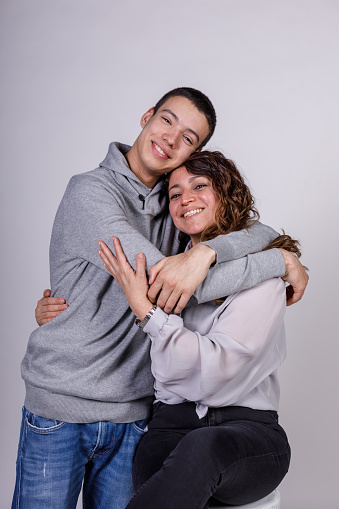Portrait of cheerful mid adult woman and her teenage son standing against white background, embracing and having bonding moments. They are looking at camera and smiling joyfully.