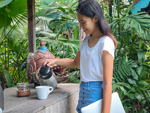 Close-up shot of smiling young Asian woman pouring hot coffee from thermos on breakfast buffet at outdoor terrace of tourist resort. She's carrying her laptop beside her.