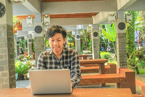 young asian man wearing flannel shirts is smiling while working remote job on grey laptop as freelancer designer, in the bench park