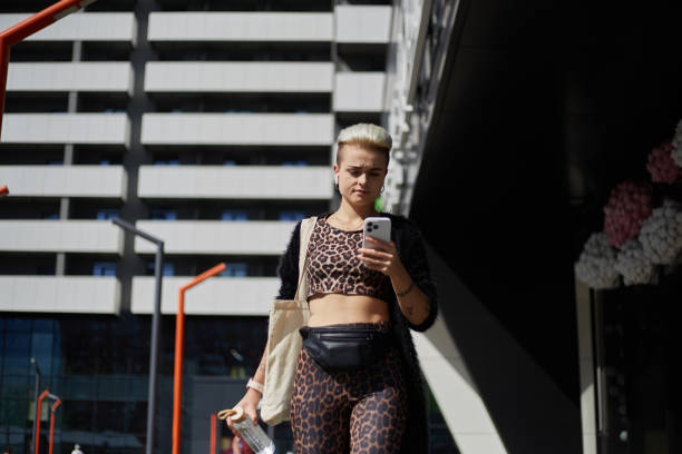 istock Young diverse female with short dyed hair walking outdoor and using a mobile app on a smartphone. Stylish white woman in leopard print clothes walks in the city center with a glass bottle of water 1491669103