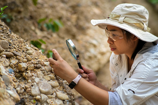 Close up female geologist using magnifying glass to examine and analyze rock, soil, sand in nature. Archaeologists explore the field. Environmental and ecology research.