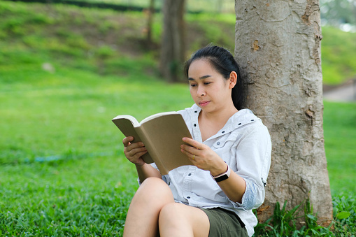 Cheerful young woman in jacket reading a book in summer park. Concentrated woman sitting on grass and studying under tree during vacation. Education concept