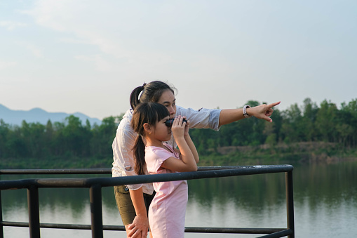 Mother teaching her little daughter to use binoculars by a reservoir in the summer garden. Explore and adventure concept.