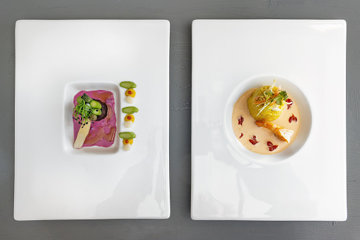 Two courses of a modern gourmet menu on white plates, asparagus in nori leaf with beetroot sauce, saffron risotto with prawn and crustacean sauce, top view from above, copy space, selected focus