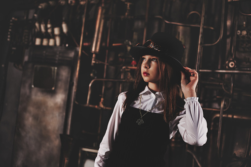 Portrait of lovely stylish teen girl in steampunk image over industry gear background, looking up away. Toned image of dreamy cute steampunk girl model actress. Adventure concept. Copy ad text space