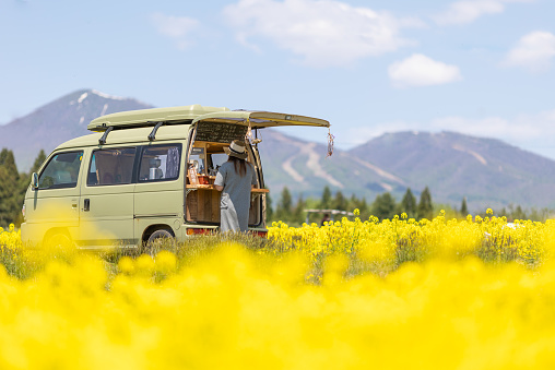 A Japanese man running a mobile coffee cafe out of his van. Parked in the countryside amongst a field of bright yellow Canola flowers. A woman is buying coffee from him.