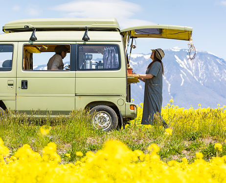 A Japanese man running a mobile coffee cafe out of his van. Parked in the countryside amongst a field of bright yellow Canola flowers. A woman is buying coffee from him.