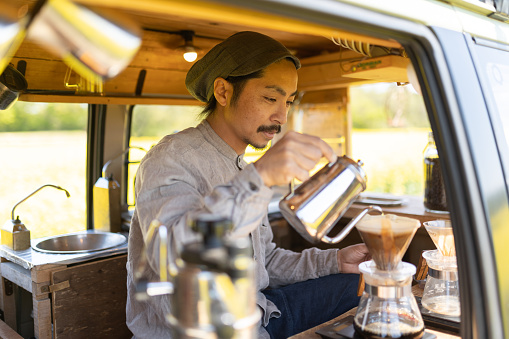 A Japanese man running a mobile coffee stand out of his van. Parked in the countryside amongst a field of bright yellow Canola flowers.