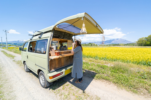 A Japanese man running a mobile coffee cafe out of his van. Parked in the countryside amongst a field of bright yellow Canola flowers. A woman is buying coffee from him.