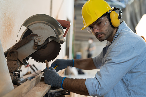 Male carpenter using electrical circular saw electric during working in wood workshop. Male joiner wearing safety uniform, gloves, helmet and working in furniture workshop