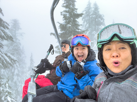 Eurasian adult daughter skier and Chinese senior mother snowboarder taking selfie on chairlift ride on a snowy winter day. Caucasian young man in background waving. North Vancouver, British Columbia, Canada.
