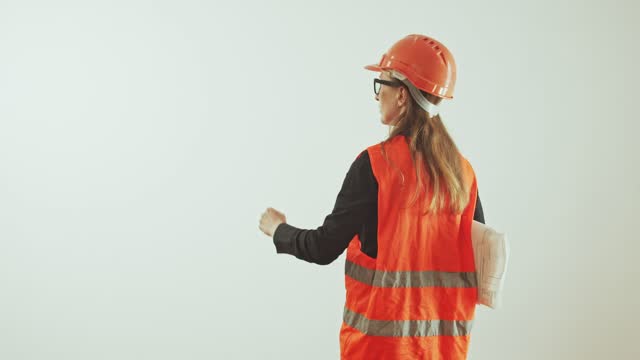 A young adult woman in glasses, a hard hat and a reflective orange vest with a stack of paper work drawings on a white background counts with her hand. Back view