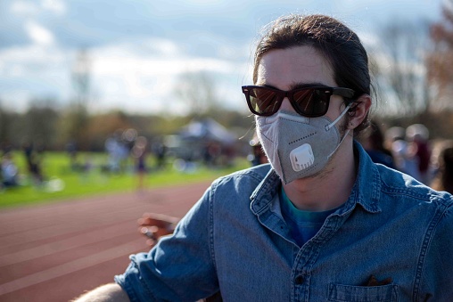 Handsome young man in a blue denim shirt stands by athletic track as a spectator. He wears a face mask and sunglasses with defocused athletic scene background. Natural light with copy space.
