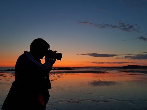 Silhouette of photographer at sunset on the beach using camera - Man taking photos in low light standing by the ocean - Concept of travel hobby and photography equipment