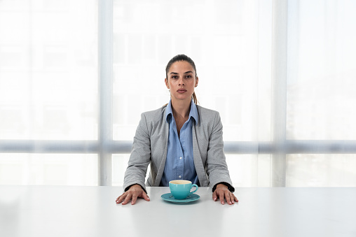 Young serious business woman in formal wear with ponytail sitting at the empty desk in the office with cup of coffee and looking at the camera. Businessperson empty face expression before work