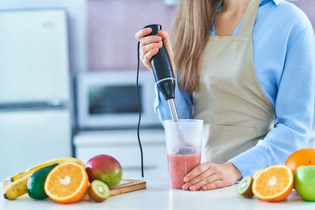 femme en tablier utilisant un mélangeur à main pour préparer un smoothie aux fruits biologiques frais à la maison dans la cuisine - batteur électrique photos et images de collection