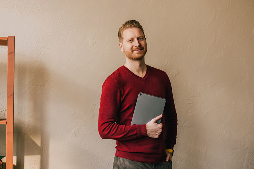 A smiling Caucasian entrepreneur looking at camera while holding his tablet. He is standing against beige wall.