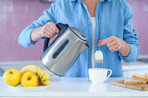 Woman putting tea bag in a cup and using an electric kettle for making tea
