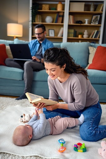 A mother reading a book to her daughter on the floor while a dad using a laptop on the sofa