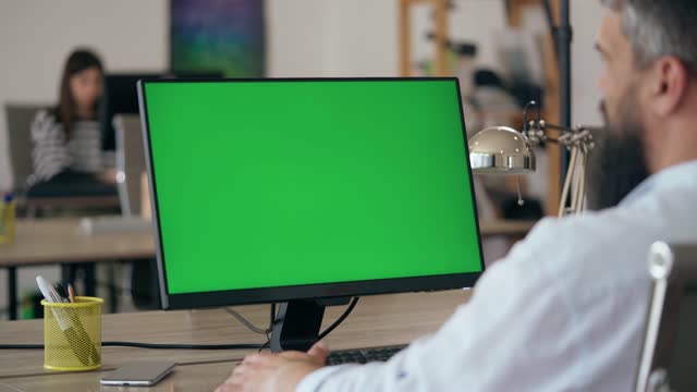 Bearded Man Uses Desktop Computer with Green Mock-up Screen In Office
