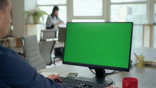 Young Man Sitting at His Desk Uses Desktop Computer with Green Mock-up Screen