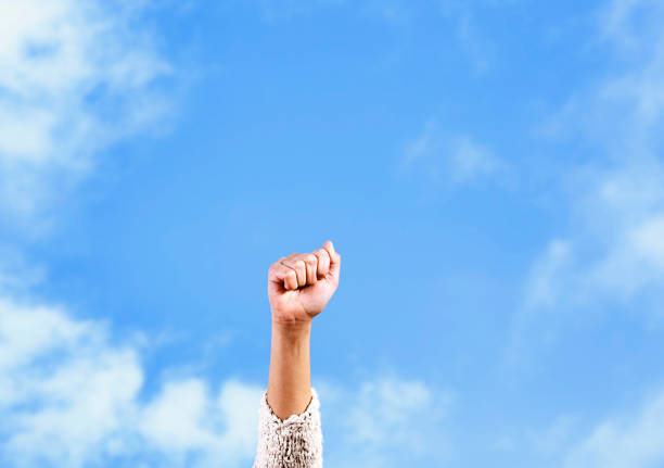 Representing inspiration and hope, a woman's clenched fist rises into clear summer sky, punching through the clouds.