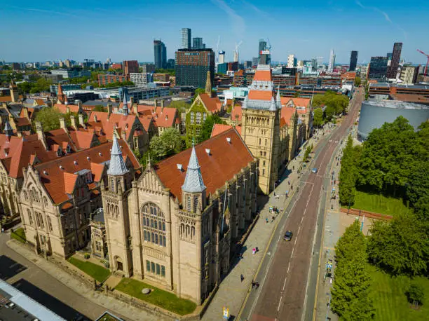 An aerial photograph of the University of Manchester on Oxford Road in Manchester, England. The photograph was taken on a bright sunny day with clear blue skies. The skyline of downtown Manchester skyline can be seen in the distance