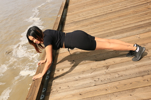 A Fijian model doing push ups as part of a workout on a floating pier on the the Fraser River. She is wearing long brown hair, a black t shirt and shorts with black shoes.