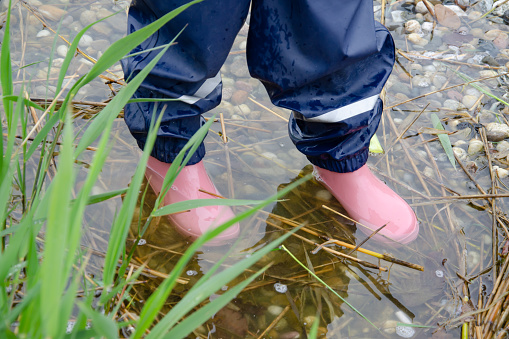 The girl's feet dressed in rubber boots and Muddy trousers.  The girl is standing in a puddle. There is grass in the foreground.
