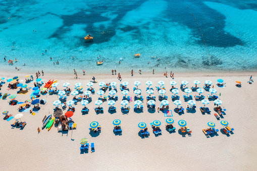 Aerial view of colorful umbrellas on sandy beach, swimming people in blue sea at summer sunny day. Sardinia, Italy. Tropical seascape. Travel and vacation. Top drone view of ocean with azure water