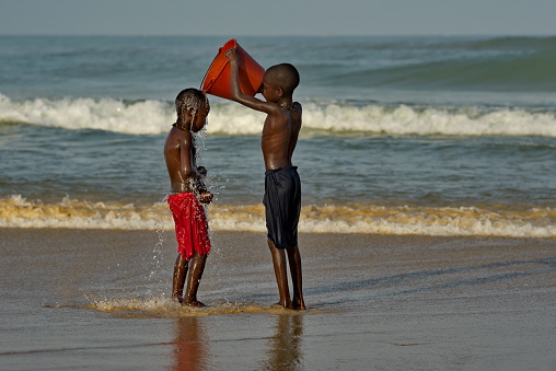 Saint-Louis. Senegal. October 11, 2021. Two village boys are doused with seawater from a plastic bucket on the sandy shore of the Atlantic Ocean.