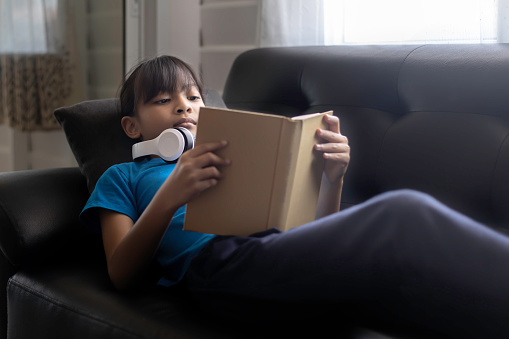 Asian Young girl lying on a black sofa reading a book in a dimly lit room.