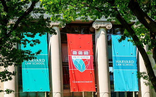 Cambridge, Massachusetts, USA - May 19, 2023: Three Harvard Law School banners hang over the entrance to Langdell Hall, at the time of graduation commencement. The red banner has the Law School Shield that has the traditional Harvard Latin motto VERITAS, meaning Truth; and under it is the latin phrase LEX ET IUSTITIA, meaning Law and Justice. Langdell Hall (c. 1905) is the largest building of Harvard Law School (HLS) of Harvard University. It is home to the school's library, the largest academic law library in the world.
