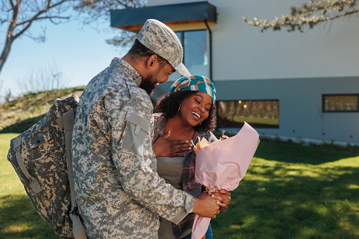 A young adult African American soldier is returning home from service and surprising his wife with a bouquet of flowers.