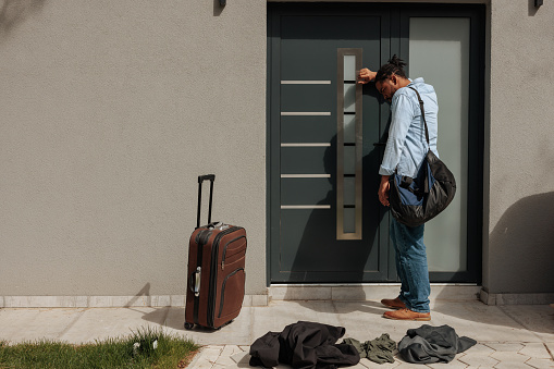 A young adult African American man is knocking on his house door with his belongings in bags and suitcases and on the floor, because his wife threw his out with copy space.