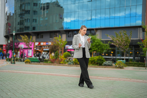 Young businesswoman holding coffee mug and looking at her phone in front of business center