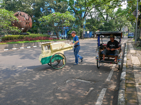 May 17, 2023 - Bandung, West Java, Indonesia.
A view of the north part of Jl. Buah Batu in the Burangrang, Lengkong district of Bandung with trees lining the road and a becak or pedicab parked on the side of the road. A man pushing a grobak food cart is crossing the road nearby.