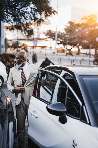 Lisbon; Vertical shot; Black male bald unshaven beard with white hairs fixing his jacket as he is about to enter his white car that is parked and adjacent to another vehicle