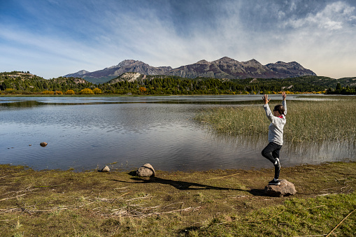 mature woman exercises on a rock in front of a lake, while contemplating the landscape of water and mountains