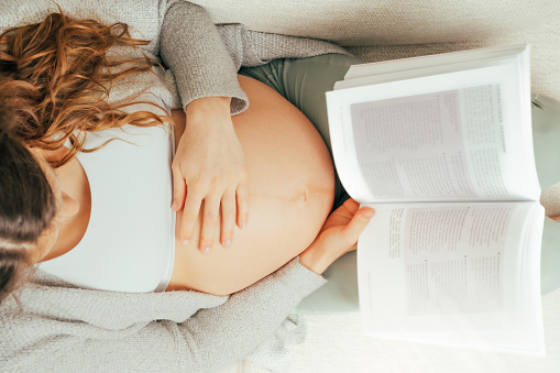 Description: Top view of a woman sitting on a sofa reading a book in the last stage of pregnancy. Pregnancy third trimester - week 34. Top view. Sunny atmosphere.