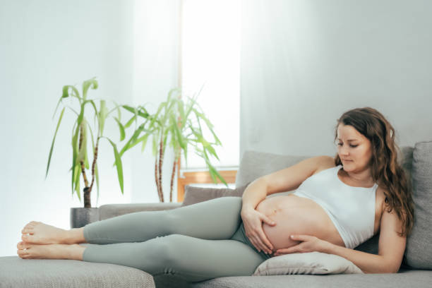 vue d’une femme allongée sur le côté sur un canapé tenant fièrement son ventre dans la dernière étape de la grossesse. grossesse troisième trimestre - semaine 34. vue latérale. fond plat blanc. - table chair white curve photos et images de collection