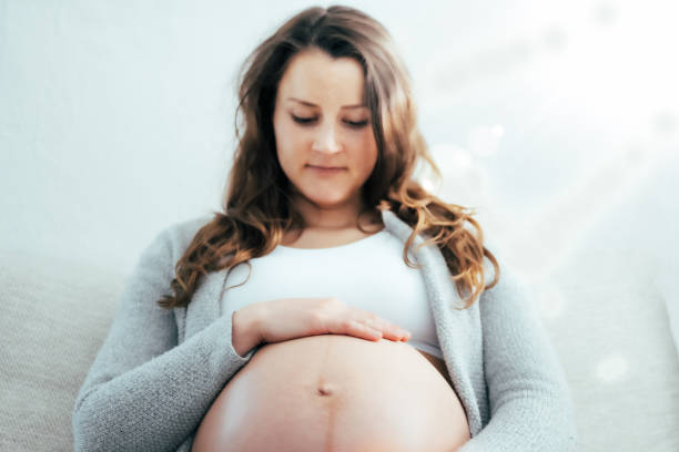 vue frontale d’une femme heureuse assise sur un canapé tenant doucement son ventre dans l’attente d’un bébé au dernier stade de la grossesse. grossesse troisième trimestre - semaine 34. vue de face. fond blanc. - table chair white curve photos et images de collection