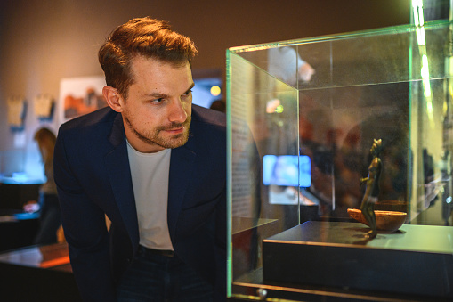 Close up of a handsome Caucasian man taking a better look at the exhibited statue in a glass case. Waist up image, his face in foreground, looking away. Reflection of lights in the glass, partly shot through glass.